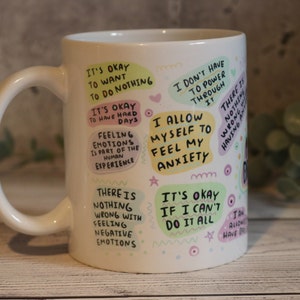 Pu&ograve; includere: Tazza in ceramica bianca con testo e illustrazioni colorate. Il testo include frasi come "Va bene voler non fare niente", "Mi permetto di sentire la mia ansia" e "Non c'&egrave; niente di sbagliato nel provare emozioni negative".