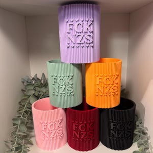 Könnte beinhalten: Eine Sammlung von sechs zylindrischen, gerippten Stiftehaltern in Lavendel, Salbeigrün, Orange, Hellrosa, Dunkelrot und Schwarz. Jeder Halter zeigt den geprägten Text FCK NZS, umgeben von kleinen Herzsymbolen auf der Vorderseite.