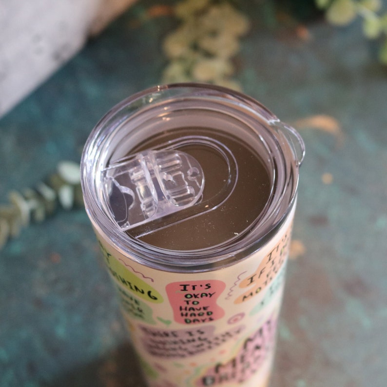 May include: A stainless steel tumbler with a clear lid and a motivational quote design. The tumbler features a variety of colorful text in a playful font, including "It's okay to have bad days".