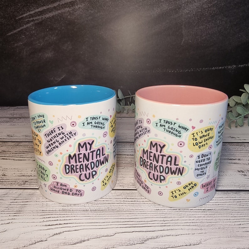 Pu&ograve; includere: Due tazze in ceramica bianca con un bordo blu e rosa. Entrambe le tazze hanno il testo "My Mental Breakdown Cup" e sono decorate con cerchi colorati e testo. Il testo sulle tazze include frasi come "Non c'&egrave; niente di sbagliato nell'avere ansia", "Mi fido di quello che sto attraversando", e "Va bene avere giornate con poca energia".