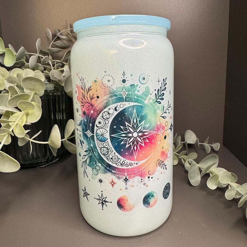 Op de afbeelding: Een witte glitterbeker met een blauwe deksel en een kleurrijk aquarelontwerp met een halve maan, sterren en bloemmotieven.