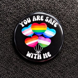 Könnte beinhalten: Ein runder schwarzer Ansteckbutton mit dem weißen Text "YOU ARE SAFE WITH ME", der über und unter einer Gruppe von fünf herzförmigen Luftballons gebogen ist. Die Ballons zeigen verschiedene Pride-Flaggen-Farbmuster, darunter Regenbogen und Transgender.