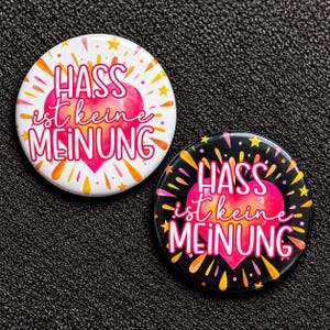 Könnte beinhalten: Zwei runde Ansteckbuttons mit einem rosa Herzmotiv und dem deutschen Text HASS ist keine MEINUNG. Ein Button hat einen weißen Hintergrund mit gelben und orangen Akzenten, der andere einen schwarzen Hintergrund mit bunten Sternen und Tropfenformen.