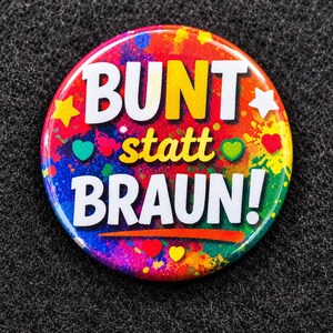 Könnte beinhalten: Ein runder Ansteckbutton mit einem lebendigen, mehrfarbigen Farbspritzer-Hintergrund. Der Text BUNT statt BRAUN! ist in großen weißen und gelben Buchstaben aufgedruckt. Kleine weiße Sterne sowie rote, grüne und gelbe Herzen sind auf dem Design verteilt.