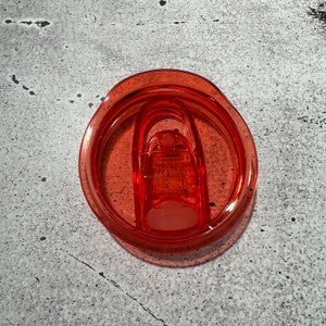 May include: A close-up of a bright orange plastic lid for a travel mug. The lid has a silicone seal and a flip-top lid.