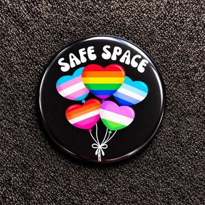 Könnte beinhalten: Ein runder schwarzer Ansteckbutton mit dem weißen Text SAFE SPACE oben. Unter dem Text befinden sich fünf herzförmige Luftballons in verschiedenen Pride-Flaggenfarben, darunter Regenbogen und Transgender, die mit weißen Schnüren zusammengebunden sind.