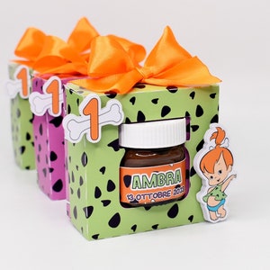 Può includere: Tre piccole scatole regalo verdi e nere con fiocchi di nastro arancioni. Le scatole hanno un osso di cartone animato e il numero uno su di esse. La scatola centrale ha un piccolo barattolo di vetro con una sostanza marrone al suo interno. Il barattolo ha un'etichetta bianca con il testo "AMBRA 13 OTTOBRE 2023". Una ragazza di cartone animato con i capelli arancioni è sul lato della scatola.