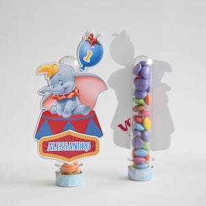 Può includere: Decorazione per feste a tema Dumbo con un elefante dei cartoni animati su un palco da circo con il nome "Alessandro". È presente anche un tubo trasparente pieno di caramelle colorate, insieme a un palloncino blu.