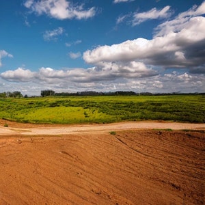 May include: A wide, open field with a dirt track leading to a grassy area. The sky is blue with white clouds.