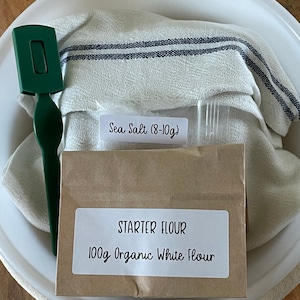May include: A white bowl with a brown paper bag containing 100g of organic white flour and a small bag of sea salt (8-10g). A white tea towel with blue stripes is in the bowl. A green spatula is also in the bowl.