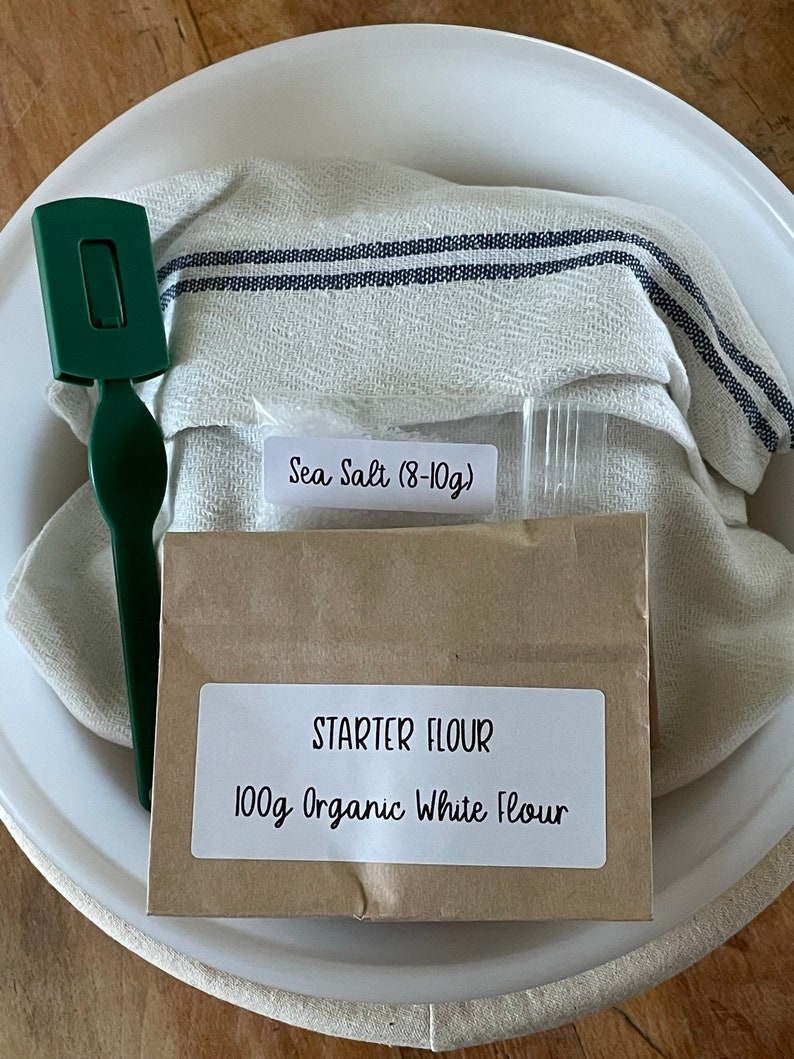May include: A white bowl with a brown paper bag containing 100g of organic white flour and a small bag of sea salt (8-10g). A white tea towel with blue stripes is in the bowl. A green spatula is also in the bowl.