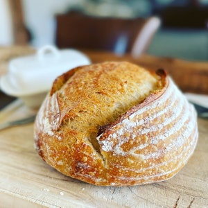 May include: A freshly baked loaf of sourdough bread with a golden brown crust and a slightly cracked surface. The bread has a rustic appearance and is sitting on a wooden cutting board.