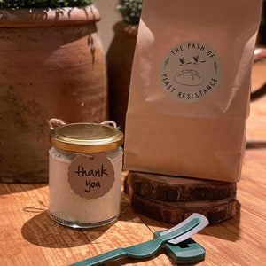 May include: A small glass jar with a gold lid and a "Thank You" tag, a green and silver bread lame, and a brown paper bag with the text "The Path of Yeast Resistance" printed on it.