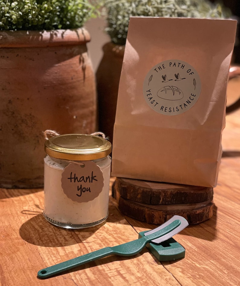 May include: A small glass jar with a gold lid and a "Thank You" tag, a green and silver bread lame, and a brown paper bag with the text "The Path of Yeast Resistance" printed on it.