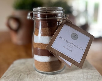 May include: A glass jar filled with layers of brown, white, and tan ingredients. A brown paper tag is attached to the jar with the text "Sourdough Brownie Mix Homemade The Original".