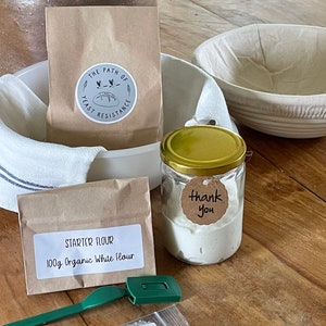 May include: A sourdough starter kit with a brown paper bag labeled "The Path of Yeast Resistance", a small brown paper bag labeled "Starter Flour 100g Organic White Flour", a glass jar with a gold lid containing sourdough starter, a small plastic bag with sea salt, a green dough scraper, and a white linen cloth.
