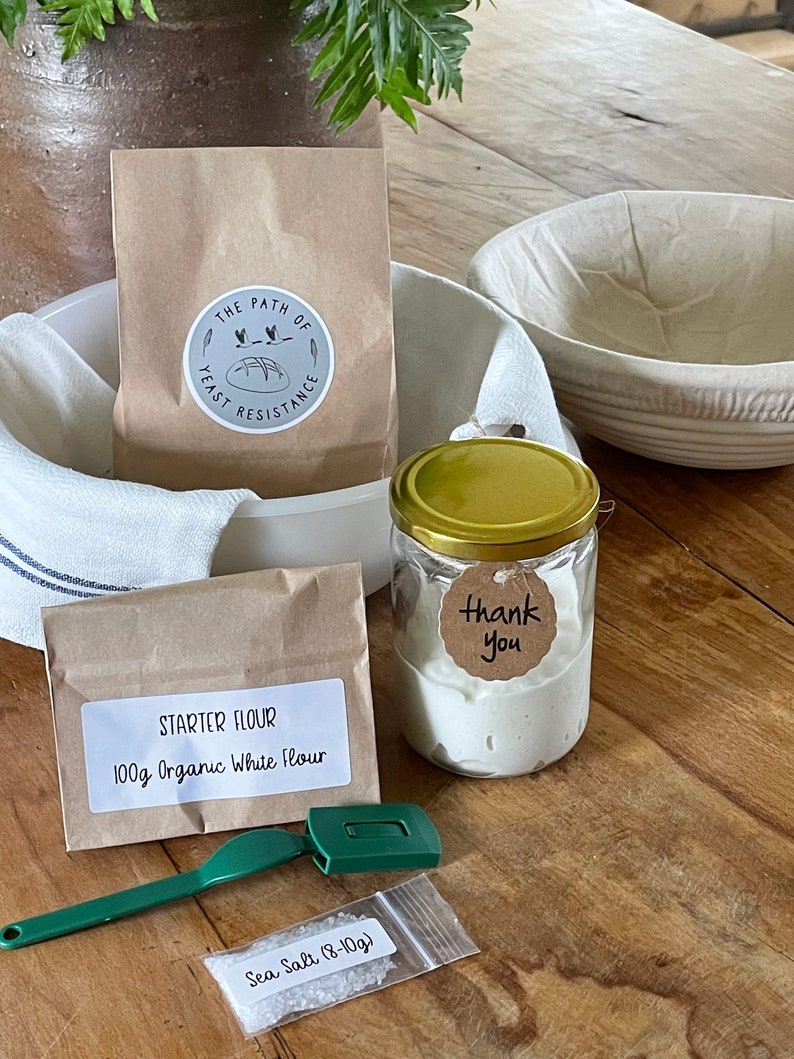 May include: A sourdough starter kit with a brown paper bag labeled "The Path of Yeast Resistance", a small brown paper bag labeled "Starter Flour 100g Organic White Flour", a glass jar with a gold lid containing sourdough starter, a small plastic bag with sea salt, a green dough scraper, and a white linen cloth.