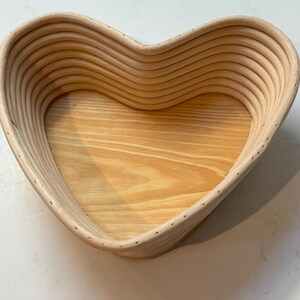 May include: A heart-shaped wooden bread proving basket with a light wood interior. The basket is made of woven strips of wood and has a smooth, natural finish.