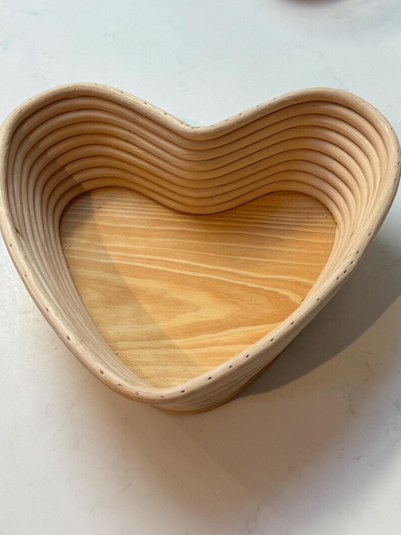 May include: A heart-shaped wooden bread proving basket with a light wood interior. The basket is made of woven strips of wood and has a smooth, natural finish.