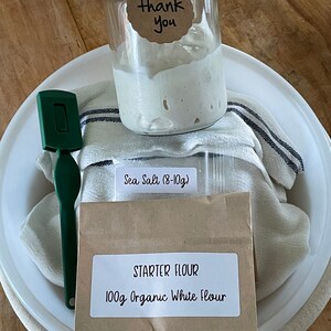 May include: A white ceramic bowl with a brown paper bag containing 100g of organic white flour and a small bag of sea salt (8-10g). A glass jar with a gold lid contains a white sourdough starter. A green spatula and a white and black striped kitchen towel are also in the bowl. The bag has a label that says "Starter Flour 100g Organic White Flour". The jar has a tag that says "Thank You".