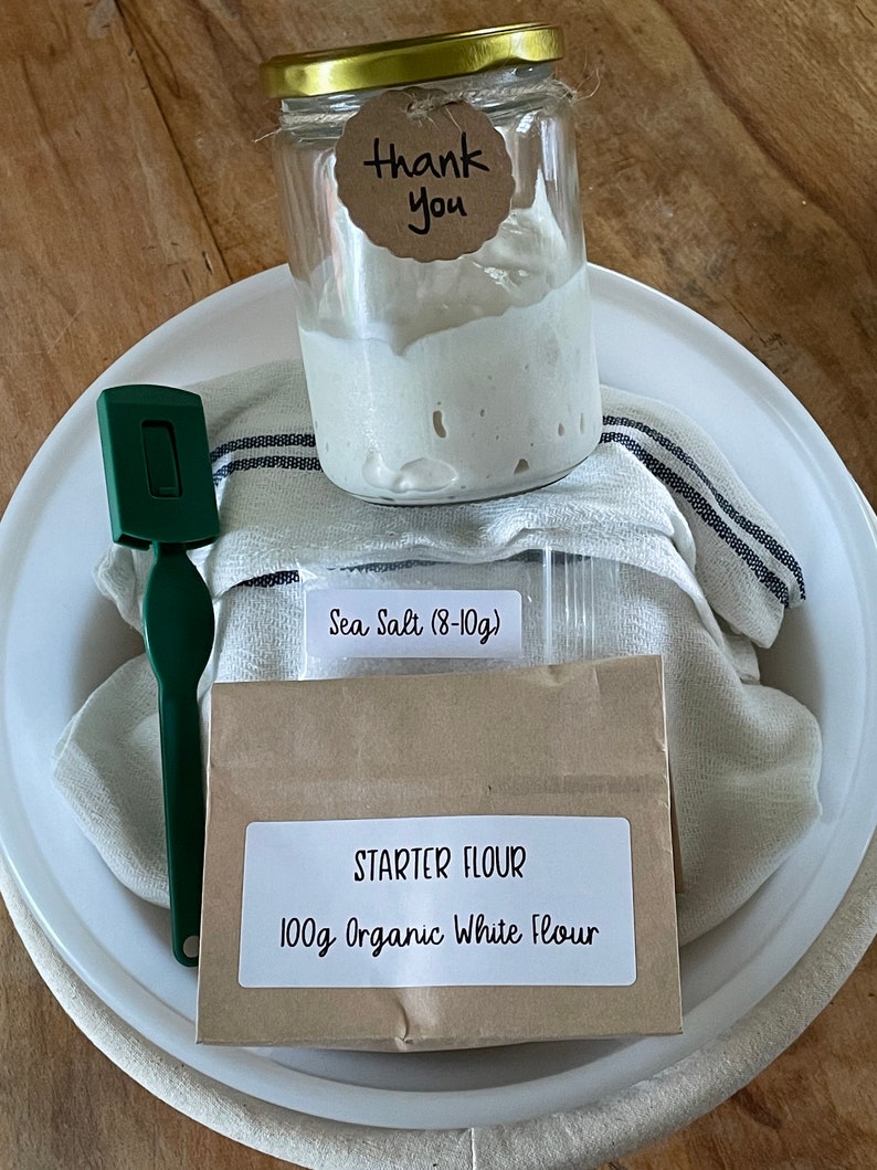 May include: A white ceramic bowl with a brown paper bag containing 100g of organic white flour and a small bag of sea salt (8-10g). A glass jar with a gold lid contains a white sourdough starter. A green spatula and a white and black striped kitchen towel are also in the bowl. The bag has a label that says "Starter Flour 100g Organic White Flour". The jar has a tag that says "Thank You".