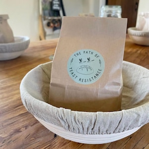 May include: A brown paper bag with a white circle sticker that says "The Path of Yeast Resistance" with a drawing of a loaf of bread and three birds flying over it. The bag is sitting in a round, light brown proofing basket with a light beige linen liner.