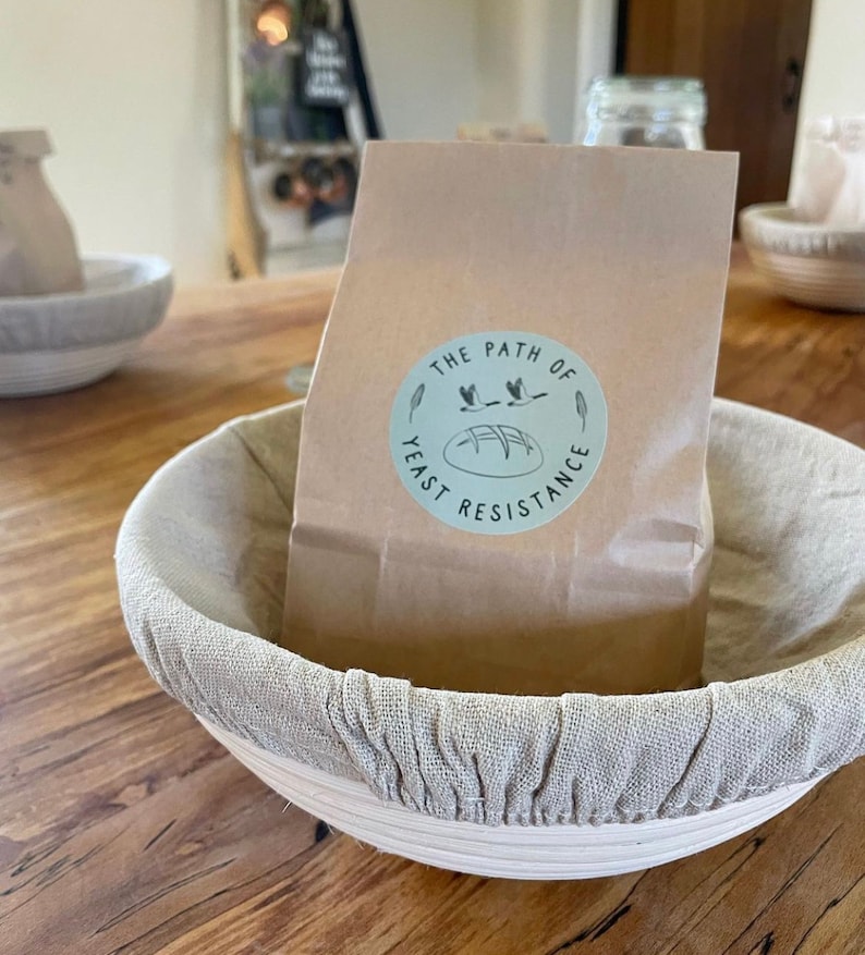 May include: A brown paper bag with a white circle sticker that says "The Path of Yeast Resistance" with a drawing of a loaf of bread and three birds flying over it. The bag is sitting in a round, light brown proofing basket with a light beige linen liner.