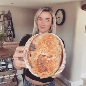 May include: A woman holds a large, round loaf of sourdough bread with a golden brown crust. The bread has a slightly irregular shape and a deep, rustic look.
