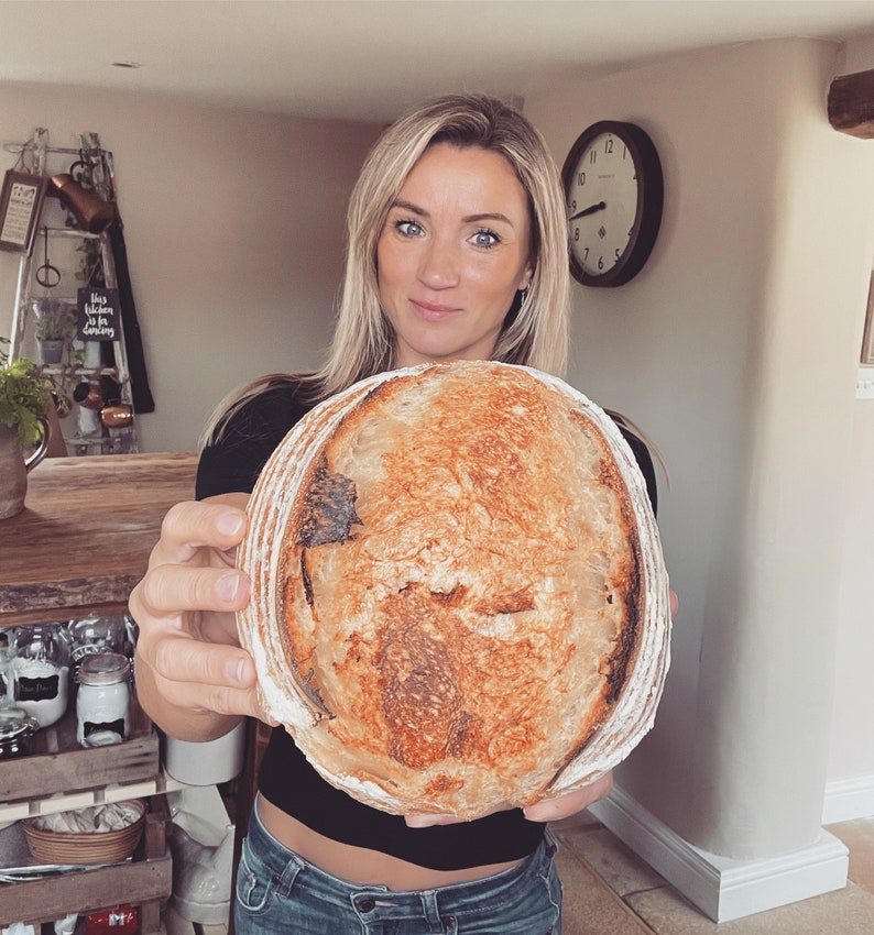 May include: A woman holds a large, round loaf of sourdough bread with a golden brown crust. The bread has a slightly irregular shape and a deep, rustic look.