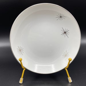 Peut inclure: Bol en céramique blanche avec un bord argenté et quatre motifs d'étoiles argentées. Le bol est sur un support jaune.