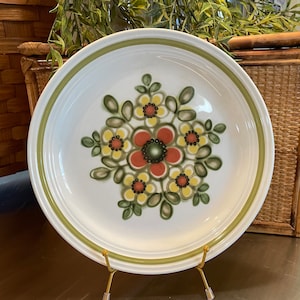 May include: A white ceramic dinner plate with a green rim and a floral design in shades of red, yellow, and green. The plate is on a gold metal stand.