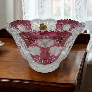 Könnte beinhalten: Eine klare Kristallschale mit gewelltem Rand und ausgestellter Form. Die Schale hat einen tiefrosa Innenraum mit weißen Blumendesigns und Blättermustern. Ein kleines Etikett ist am Rand befestigt.