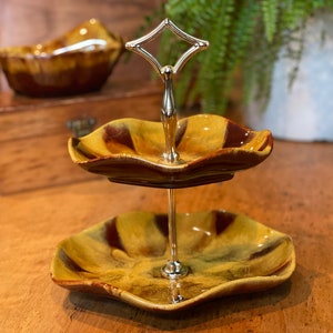 May include: Two-tier serving stand with a gold metal base and handle. The stand features two brown and yellow ceramic dishes with a wavy edge.