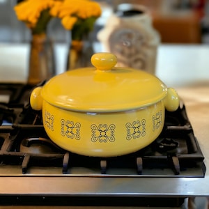 Puede incluir: Una olla de cerámica amarilla con tapa y diseño de espiral negro. La olla está sobre una estufa negra.