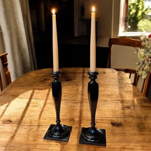 Könnte beinhalten: Zwei schwarze Kerzenhalter mit brennenden cremefarbenen Kerzen auf einem Holztisch. Die Kerzenhalter haben einen quadratischen Sockel und ein langes, schlankes Design. Der Tisch ist aus Holz mit natürlichem Finish. Der Hintergrund ist verschwommen, mit einem Fenster und einem Stuhl sichtbar.