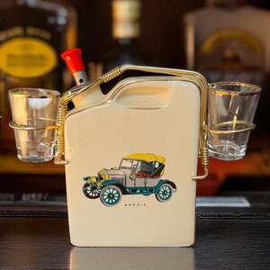 May include: Cream ceramic liquor dispenser with a vintage car illustration. It has a red cap, gold-coloured metal accents, and two shot glass holders. The car has a yellow top and teal wheels.
