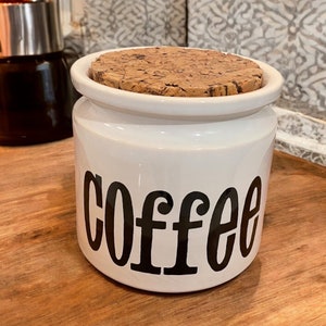 Pode incluir: Um pote de café de cerâmica branca com tampa de cortiça e a inscrição "coffee" em preto.