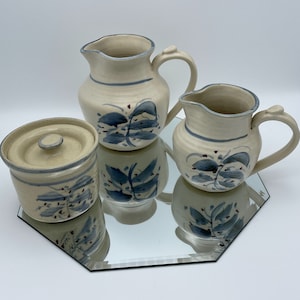 May include: A set of three ceramic kitchenware pieces with a cream base and blue and red floral designs. The set includes a creamer, a sugar bowl with a lid, and a jug.