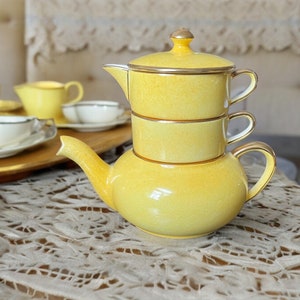 May include: A yellow ceramic teapot with a tiered design. The teapot has a gold trim and a lid with a knob.