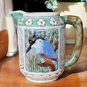 Puede incluir: Jarra de cerámica con un diseño en relieve detallado. La jarra presenta un pájaro azul posado en una rama, rodeado de follaje. El asa y el borde tienen una combinación de colores verde y marrón, con detalles florales blancos.