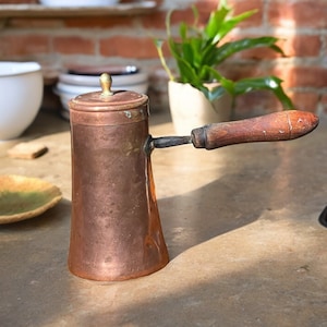 Może przedstawiać: Karafka do kawy wykonana z miedzi z drewnianym uchwytem. Karafka ma zaokrąglony korpus i dzióbek po boku. Uchwyt jest przymocowany do karafki metalową obręczą.