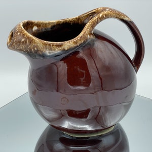 May include: A brown ceramic jug with a glossy finish. The jug has a rounded body, a curved handle, and a spout with a lighter brown rim. The jug is likely vintage and suitable for serving beverages.