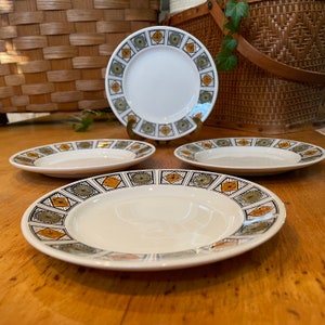 Peut inclure: Quatre assiettes en céramique blanche avec un motif géométrique dans des tons de brun, vert et orange. Les assiettes sont disposées en rangée sur une surface en bois.