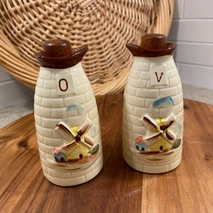 Può includere: Un set di due caraffe per olio e aceto in ceramica vintage con un motivo a mattoni e un design a mulino a vento. Le caraffe sono etichettate "O" e "V" e hanno tappi marroni.