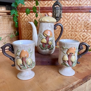 May include: A ceramic teapot and two mugs featuring mushroom designs. The teapot has a brown and white body with a yellow lid and brown handle. The mugs are brown and white with brown handles. All three pieces have a mushroom motif with brown, orange, and yellow mushrooms.