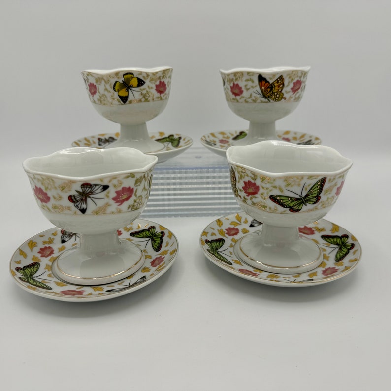 Peut inclure: Ensemble de quatre coupes &agrave; dessert en porcelaine blanche avec soucoupes assorties. Chaque coupe et soucoupe pr&eacute;sente un motif floral avec des papillons dans des tons de vert, jaune, orange et marron.