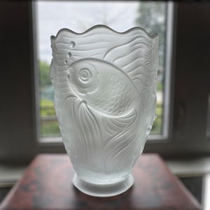 Könnte beinhalten: Eine Vase aus mattiertem Glas mit Fischdesign. Die Vase zeigt ein detailliertes Relief eines Fisches, der zwischen Wasserpflanzen und Blasen schwimmt. Die Vase hat einen wellenförmigen Rand und einen runden Sockel. Die Vase ist etwa 25 cm hoch.