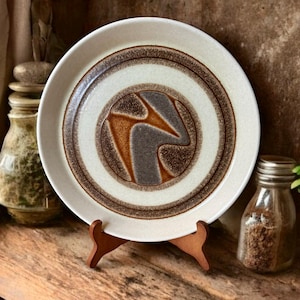 May include: A white ceramic dinner plate with a brown and orange abstract design in the center. The plate has a white rim and brown concentric circles around the center design.