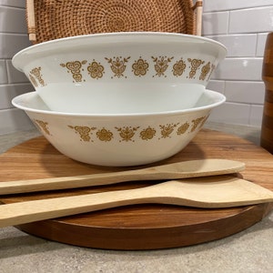 Puede incluir: Dos tazones de mezclar blancos con un patrón floral dorado, apilados uno encima del otro, sobre una superficie de madera con dos cucharas de madera.