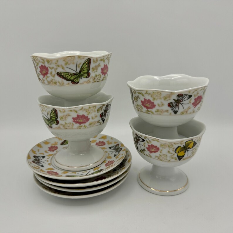 Peut inclure: Ensemble de quatre coupes &agrave; dessert en porcelaine blanche avec un pied et un motif de papillon et de fleurs. Les coupes sont empil&eacute;es par deux et se trouvent sur une surface blanche.